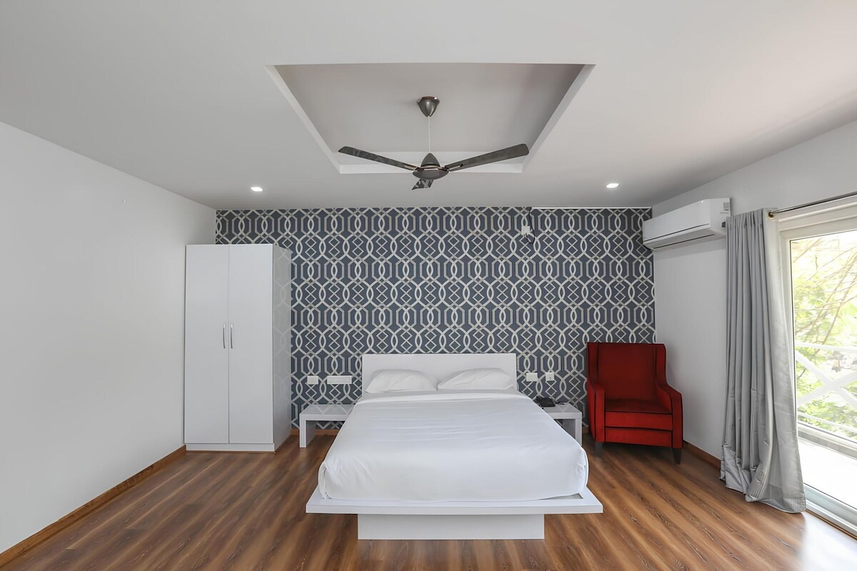 A bedroom with a bed, a red chair, a wardrobe, a ceiling fan, and patterned wallpaper.