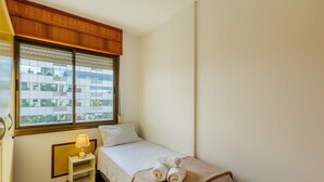Ein kleines, ordentlich eingerichtetes Schlafzimmer mit einem Bett, einem Nachttisch mit Lampe und einem Fenster mit Jalousien.