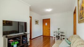 Una sala de estar con un televisor de pantalla plana sobre un pedestal de madera, un sofá, una mesa de comedor con sillas y una puerta al fondo.