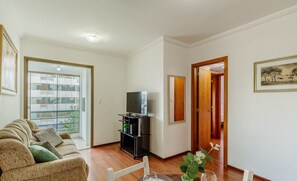 Una sala de estar con un sofá, un televisor sobre un mueble, una mesa de comedor con sillas y vistas a un balcón exterior por un ventanal.