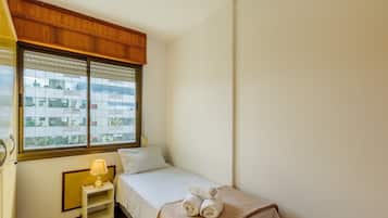 A small, neatly arranged bedroom with a bed, a bedside table with a lamp, and a window with blinds.