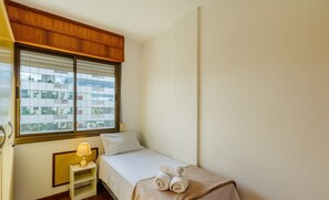 Ein kleines, ordentlich eingerichtetes Schlafzimmer mit einem Bett, einem Nachttisch mit Lampe und einem Fenster mit Jalousien.