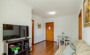 Un salon avec un téléviseur à écran plat posé sur un meuble en bois, un canapé, une table à manger avec des chaises, et une porte visible au fond.