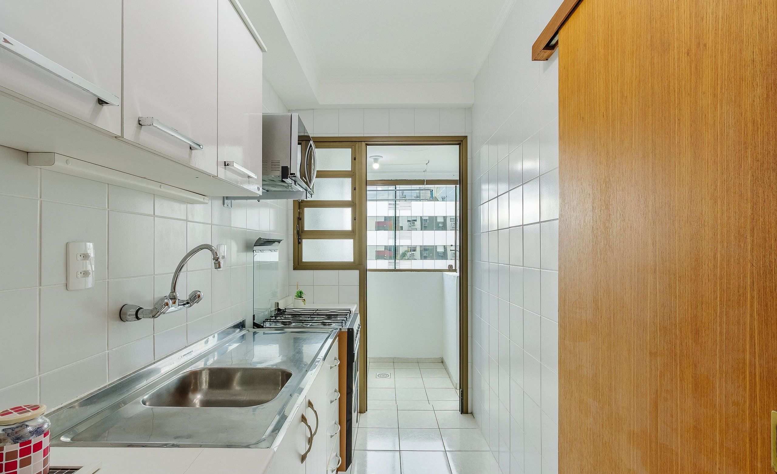 A narrow kitchen with white cabinets, a stainless steel sink, and a wooden door leading to a balcony.
