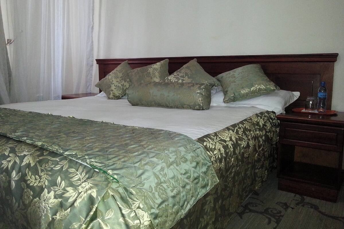 A neatly made bed with a green floral bedspread and matching pillows, a wooden headboard, a nightstand with a bottle and glass, and a white curtain.