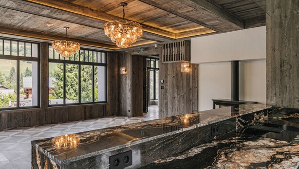 A spacious room with a large marble countertop, wooden ceiling beams, and large windows offering a view of greenery outside. - L'Écrin  (Megève)