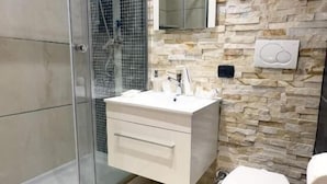 Une salle de bain moderne avec une douche à parois de verre, un meuble-lavabo blanc doté d’un miroir et un mur à l’aspect pierre.