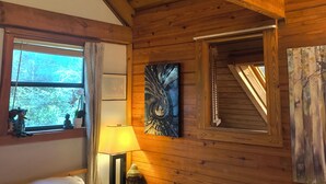 A wooden cabin room with a bed, a window with curtains, a painting, and a small Buddha statue.