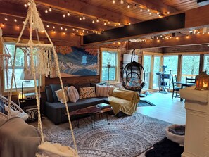 Un interior acogedor de cabaña con techo de madera, una silla colgante de macramé, un sofá con cojines, una alfombra grande y un cuadro de montañas.