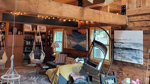 A cozy cabin interior with a wooden ceiling, a ceiling fan, a dining table with chairs, a hanging chair, a bookshelf, and a painting on the wall.