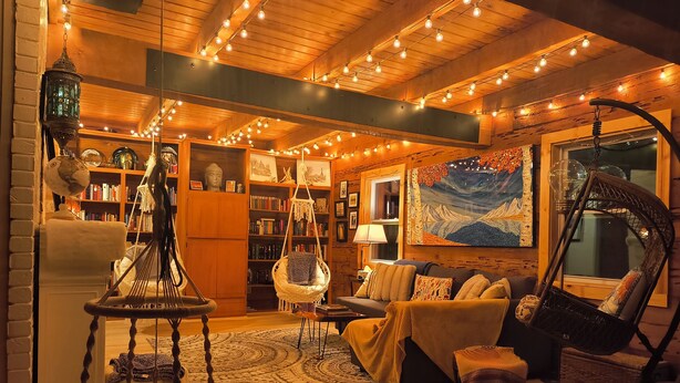 A cozy living room with a wooden ceiling, string lights, a hanging chair, a sofa, and a bookshelf.