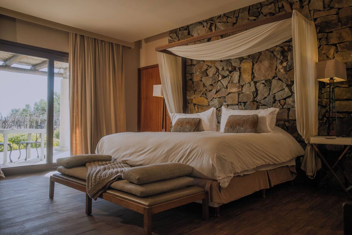 A bedroom with a stone wall, a canopy bed, a bench, and a view through a glass door.