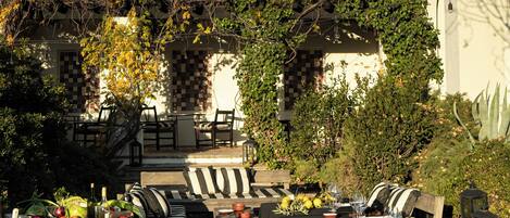 Un área de comedor al aire libre con un banco a cuadros, una mesa dispuesta con copas de vino y vistas a un edificio con tejado de azulejos e hiedra.