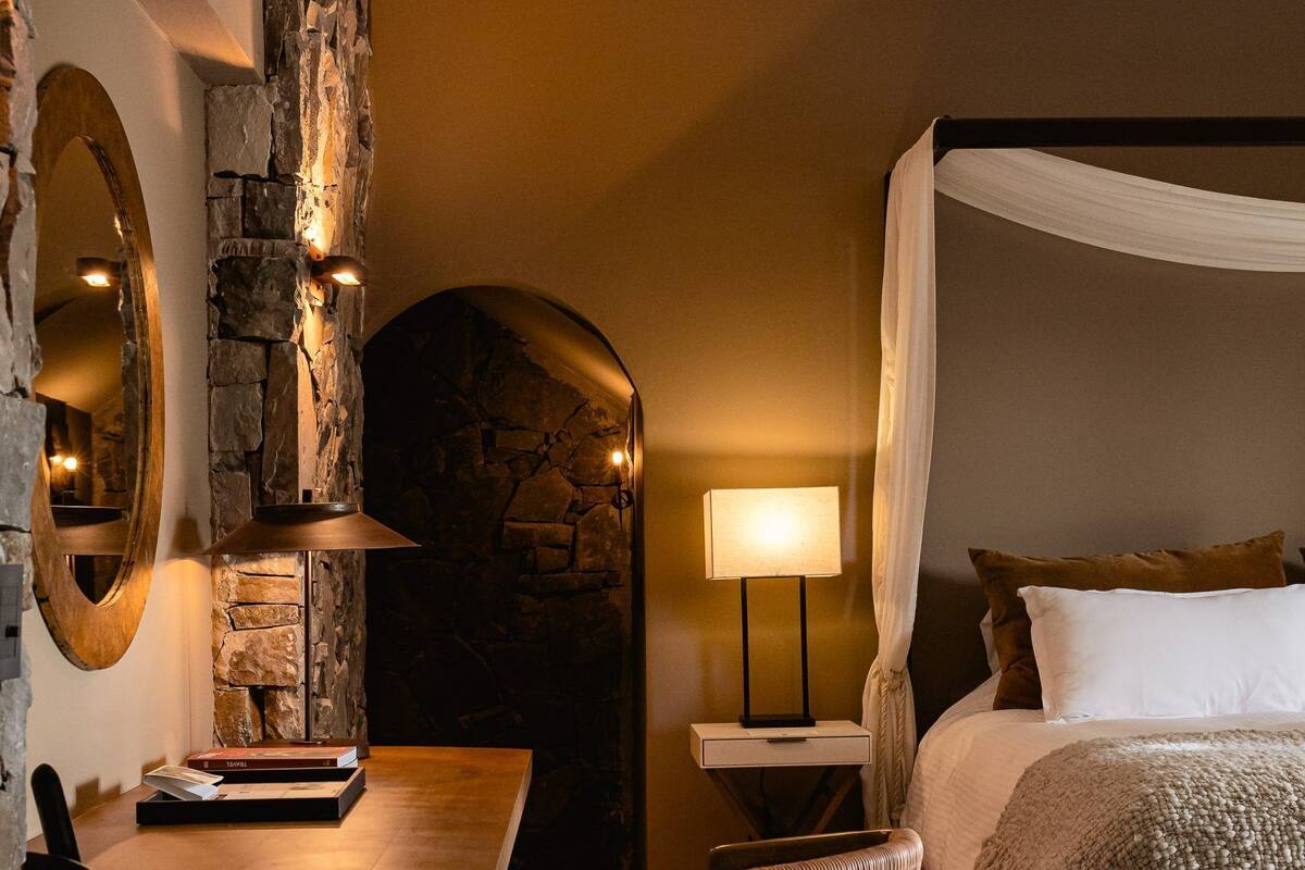 A bedroom with a stone accent wall, a canopy bed, a desk with a chair, and a bedside lamp.