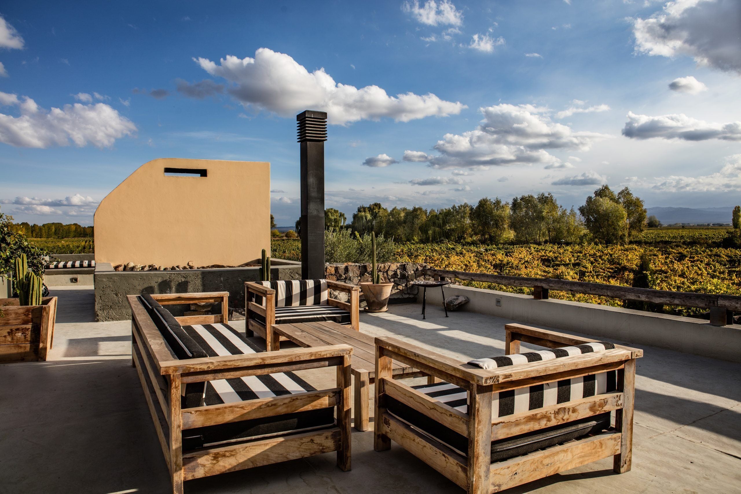 A rooftop terrace with wooden furniture, a large planter, and a tall chimney.