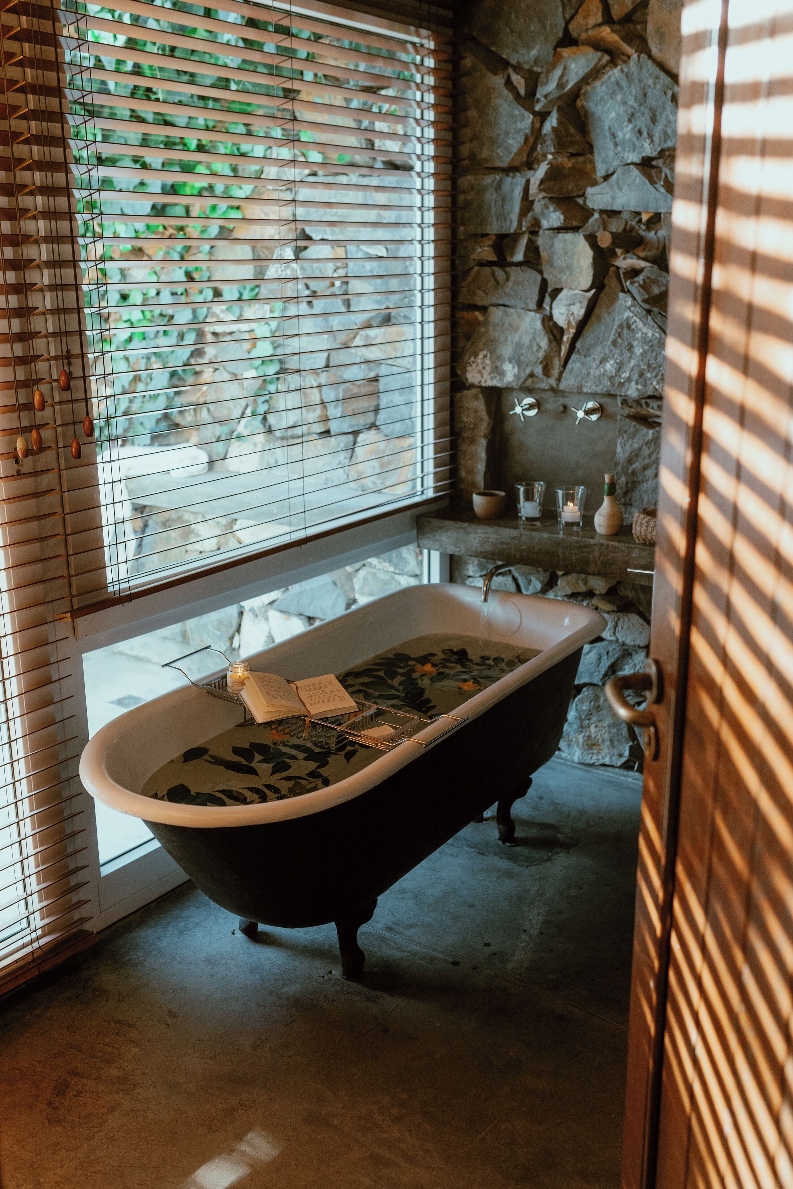 A bathroom with a freestanding bathtub, stone wall, and a window with blinds.