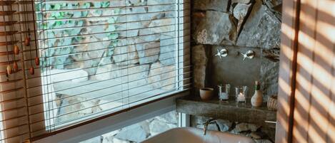 A bathroom with a freestanding bathtub, stone wall, and a window with blinds.