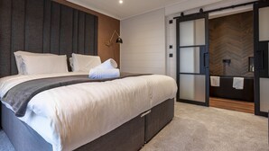 Ein modernes Schlafzimmer mit einem großen Bett, einem dunklen Kopfteil, einer Schiebetür zu einem Badezimmer und einem Fischgrätparkettboden.