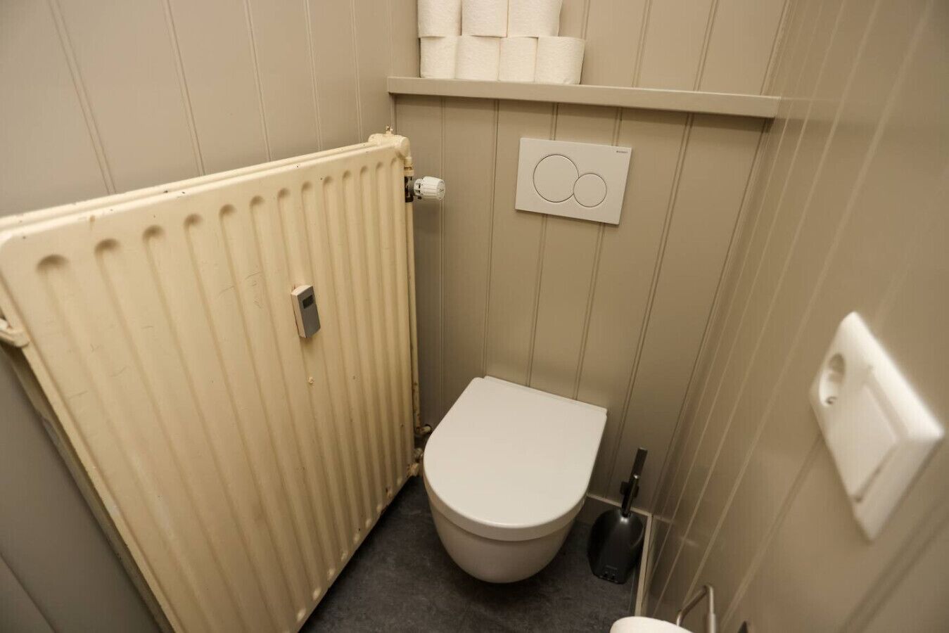A small, compact bathroom with a toilet, a radiator, and a shelf with towels.