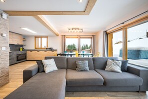 A modern living room with a grey sectional sofa, wooden beams, a kitchen with dark cabinets, and a dining area with yellow chairs.