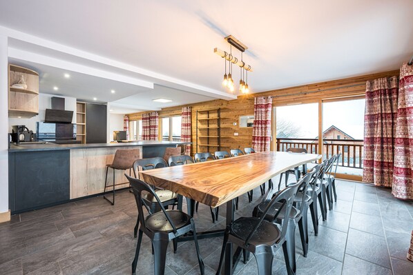 Une cuisine moderne avec un îlot central en bois, un coin repas doté d’une grande table et de chaises, et de grandes fenêtres agrémentées de rideaux.