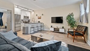 A modern living room with a sofa, coffee table, and a TV. There is a kitchen area with a microwave, refrigerator, and bar stools. - Cozy King Bed Suite ~ Free Parking & Ground Floor! (Pittsburgh)