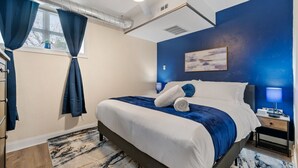 A bedroom with a bed, a nightstand, a lamp, a window with curtains, and a painting on the wall. - Cozy King Bed Suite ~ Free Parking & Ground Floor! (Pittsburgh)