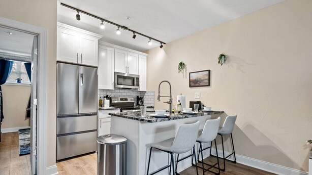 A modern kitchen with white cabinets, stainless steel appliances, a marble island with bar stools, and a view of the outdoors. - Cozy King Bed Suite ~ Free Parking & Ground Floor! (Pittsburgh)