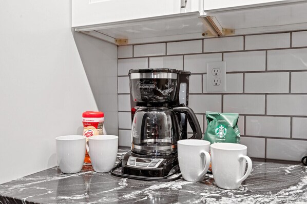 Una encimera de cocina con una cafetera, dos tazas blancas, un tarro de leche para café y una bolsa de Starbucks.