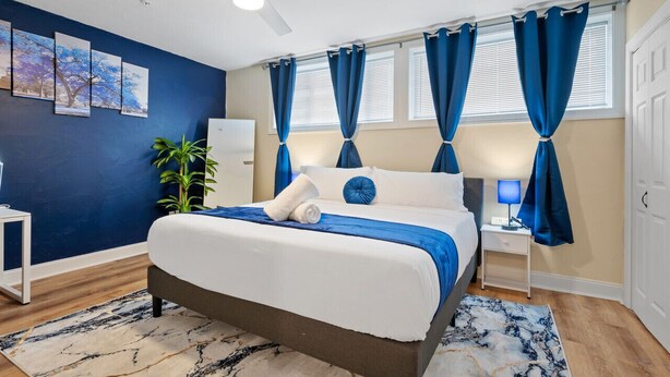 Un dormitorio con cama, ventilador de techo, mesita de noche con lámpara, ventana con cortinas y pared con cuadros enmarcados.