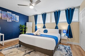 Un dormitorio con cama, ventilador de techo, mesita de noche con lámpara, ventana con cortinas y pared con cuadros enmarcados.