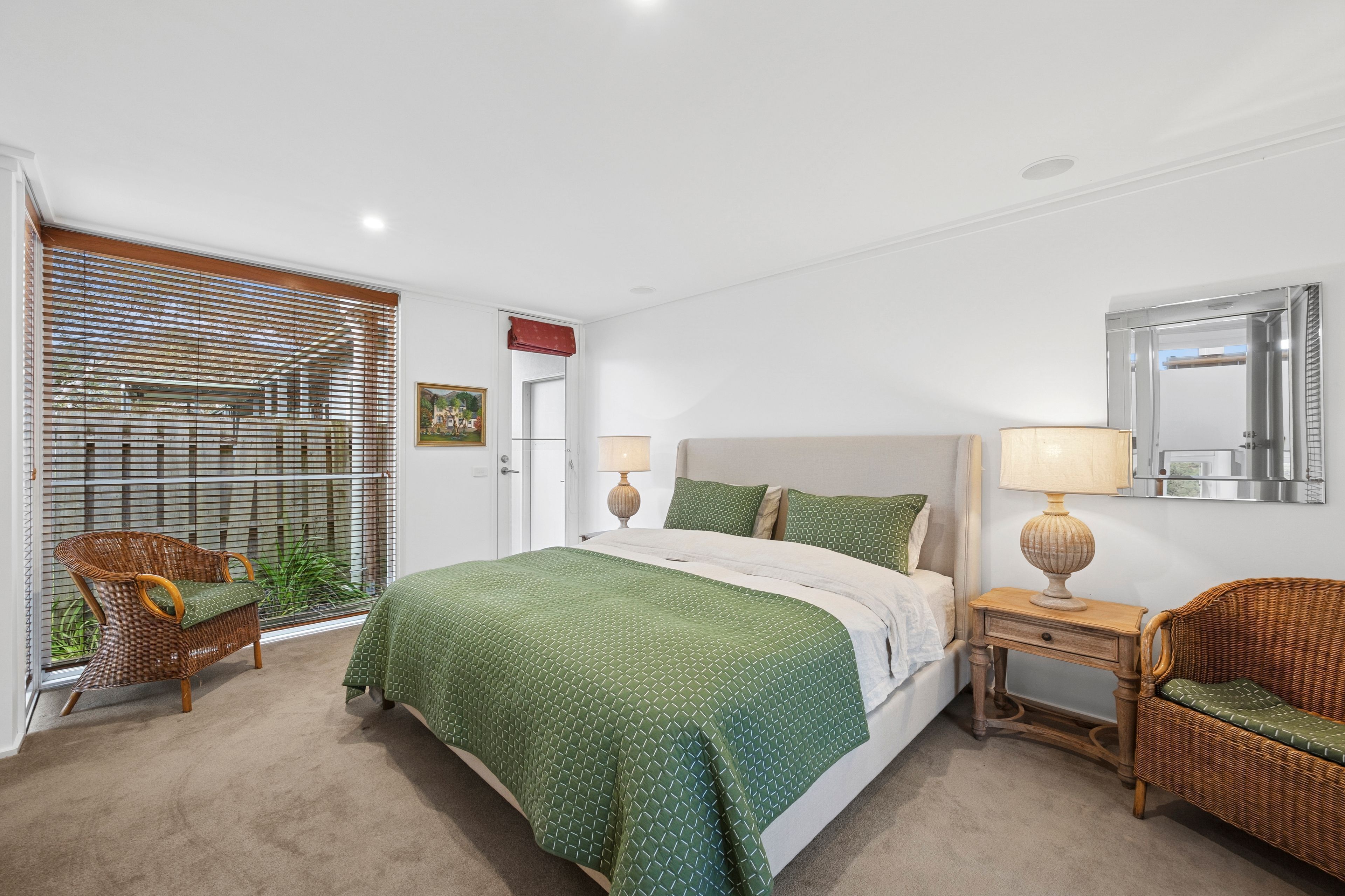 A bedroom with a bed, two bedside tables, a chair, a window with blinds, and a mirror on the wall.