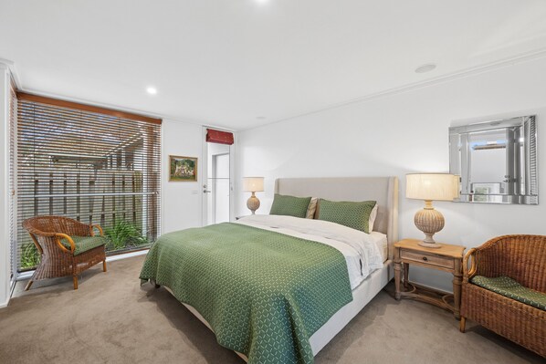 A bedroom with a bed, two bedside tables, a chair, a window with blinds, and a mirror on the wall. - Coastal Style (Lorne)