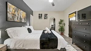 A modern bedroom with a bed, a ceiling fan, a large abstract painting, a dresser, and a potted plant.
