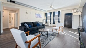 A modern living room with a dark grey sofa, white armchairs, and a coffee table. There is a ceiling fan, a large window with curtains, and a door with a dark frame. - Bright & Upscale|King Bed|Free Parking w/Workspace (Pittsburgh)
