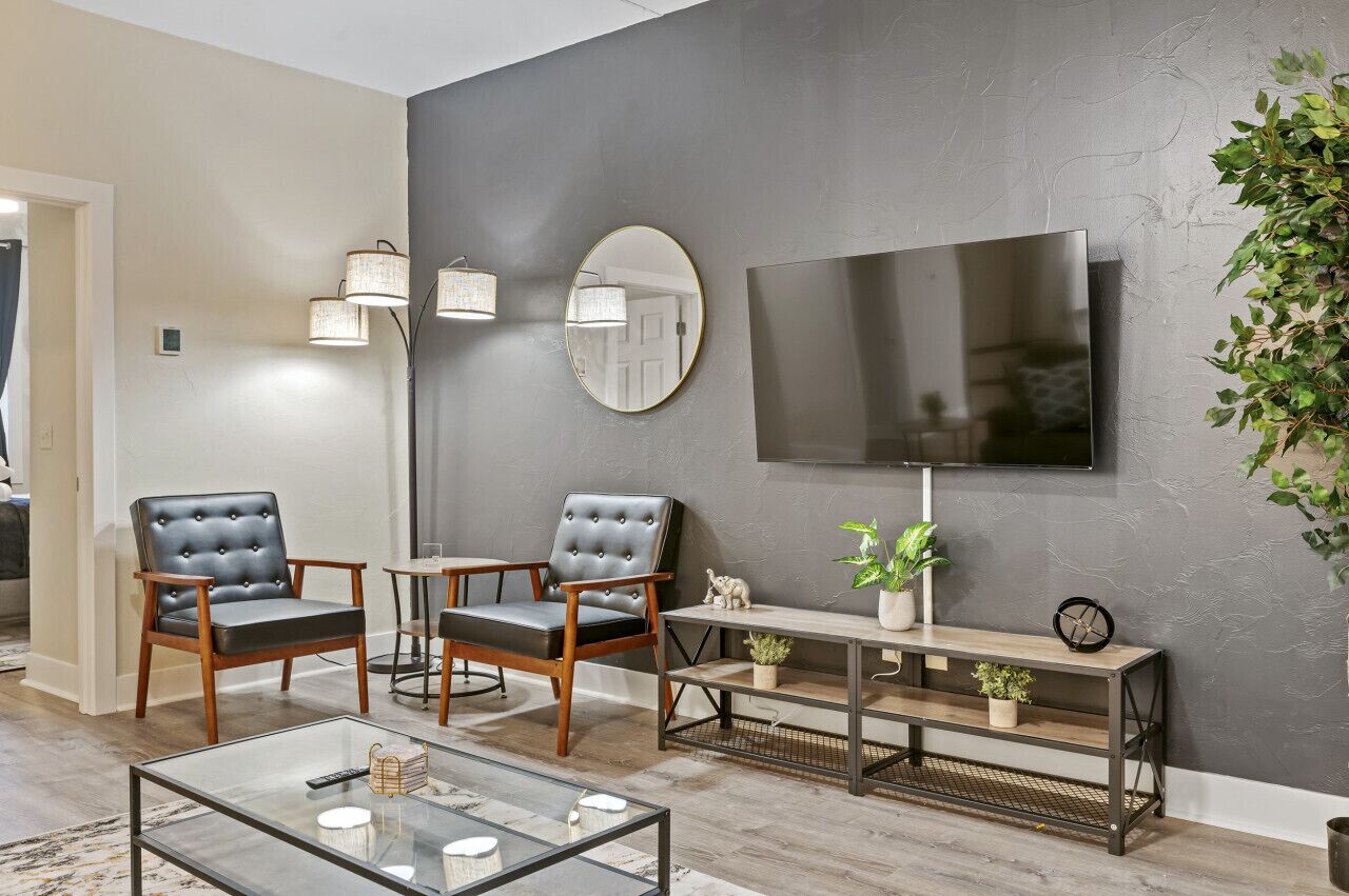 A modern living room with two leather armchairs, a glass coffee table, a TV mounted on a grey wall, and a small plant on a wooden console table.