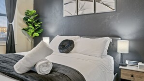 Un lit bien fait, avec du linge de lit blanc et une couverture sombre, une table de chevet avec une lampe, et une œuvre d’art accrochée au mur au-dessus du lit.