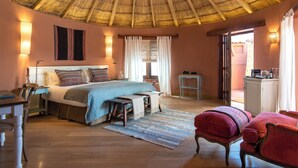 Une chambre avec un toit de chaume, un lit recouvert d’une couverture bleue, un bureau avec une chaise et une petite table avec une lampe.