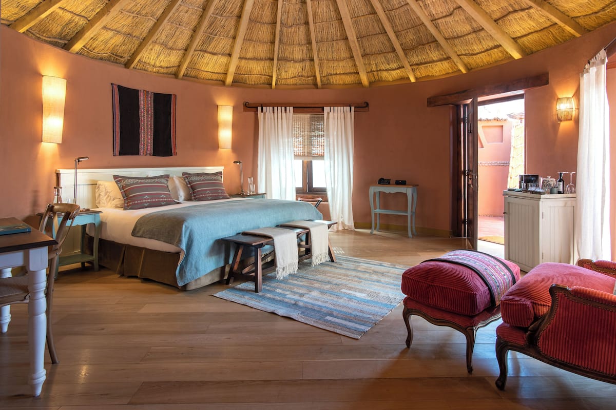 Une chambre avec un toit de chaume, un lit recouvert d’une couverture bleue, un bureau avec une chaise et une petite table avec une lampe.