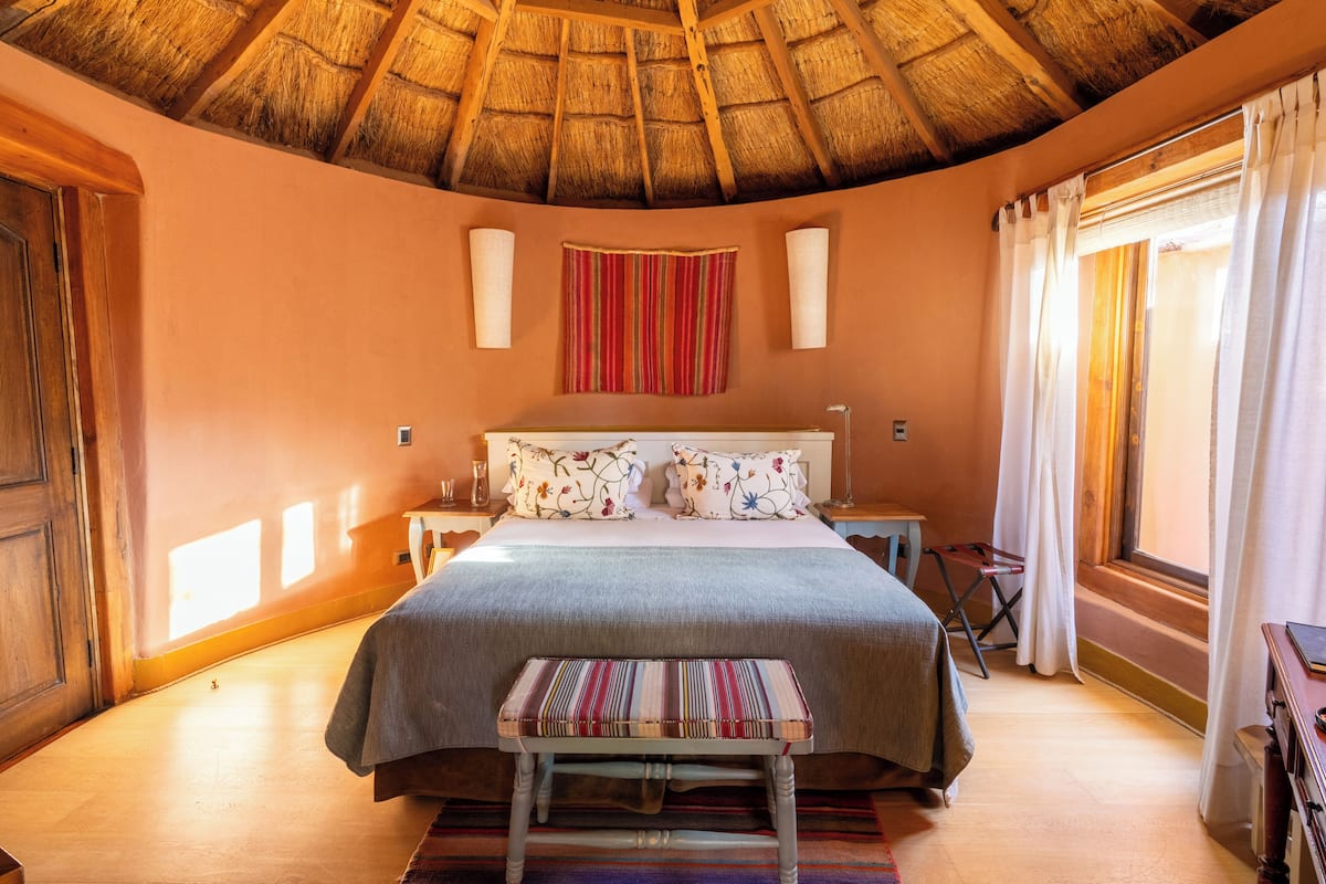 Une chambre avec un toit de chaume, un lit agrémenté d’oreillers à motifs floraux, un banc et une fenêtre avec des rideaux.