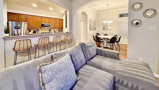 A modern living room with a grey sofa, a dining area with a table and chairs, and a kitchen with wooden cabinets and a refrigerator. - Gorgeous house in The Woodlands (Spring)