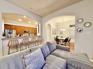 A modern living room with a grey sofa, a dining area with a table and chairs, and a kitchen with wooden cabinets and a refrigerator. - Gorgeous house in The Woodlands (Spring)