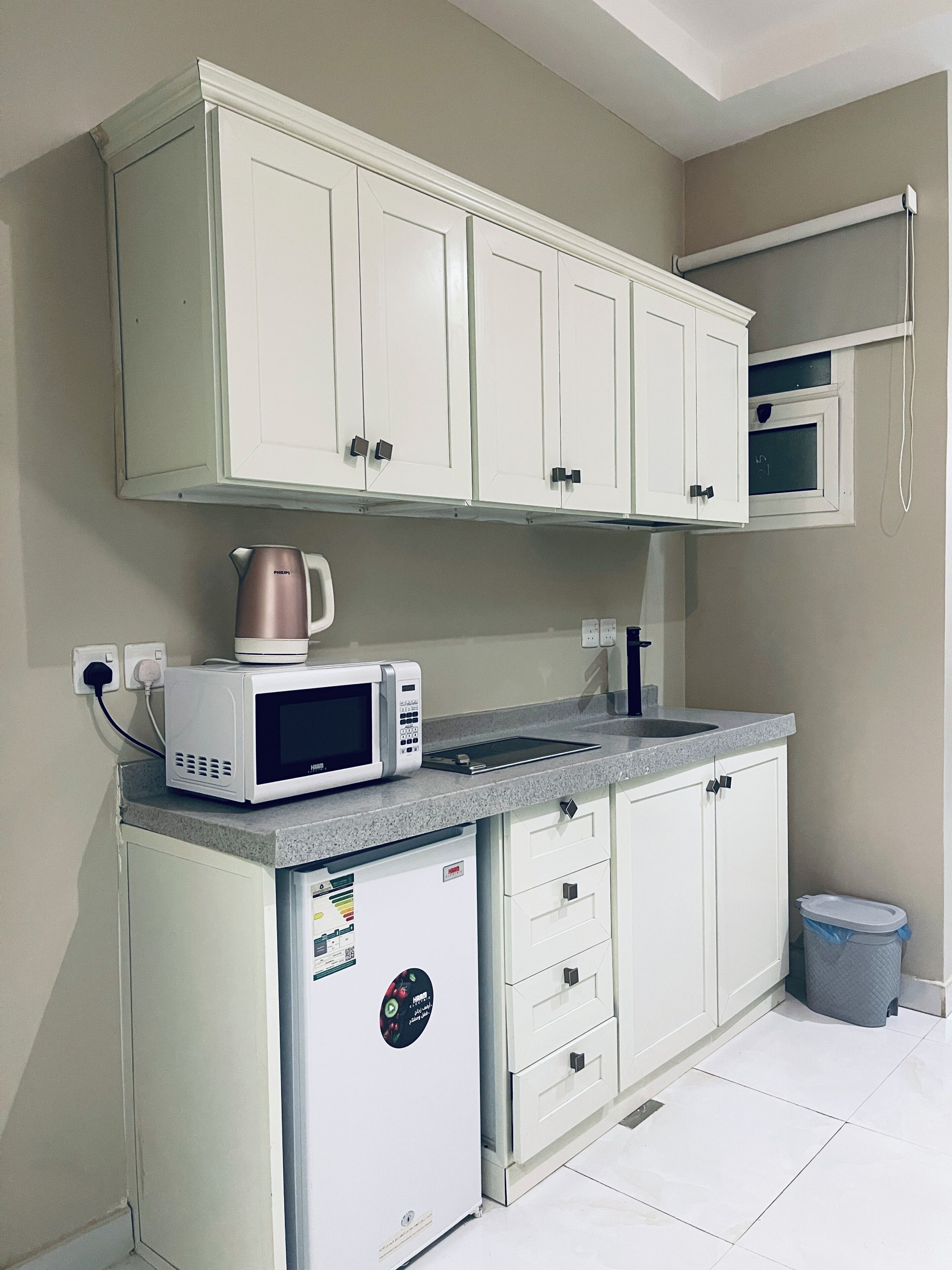 Una cocina con gabinetes blancos, microondas, hervidor y cubo de basura.