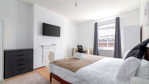 A modern hotel room with a large bed, a flat-screen TV mounted on the wall, a desk with a chair, and a window with curtains. - Spacious apartment in London (London)