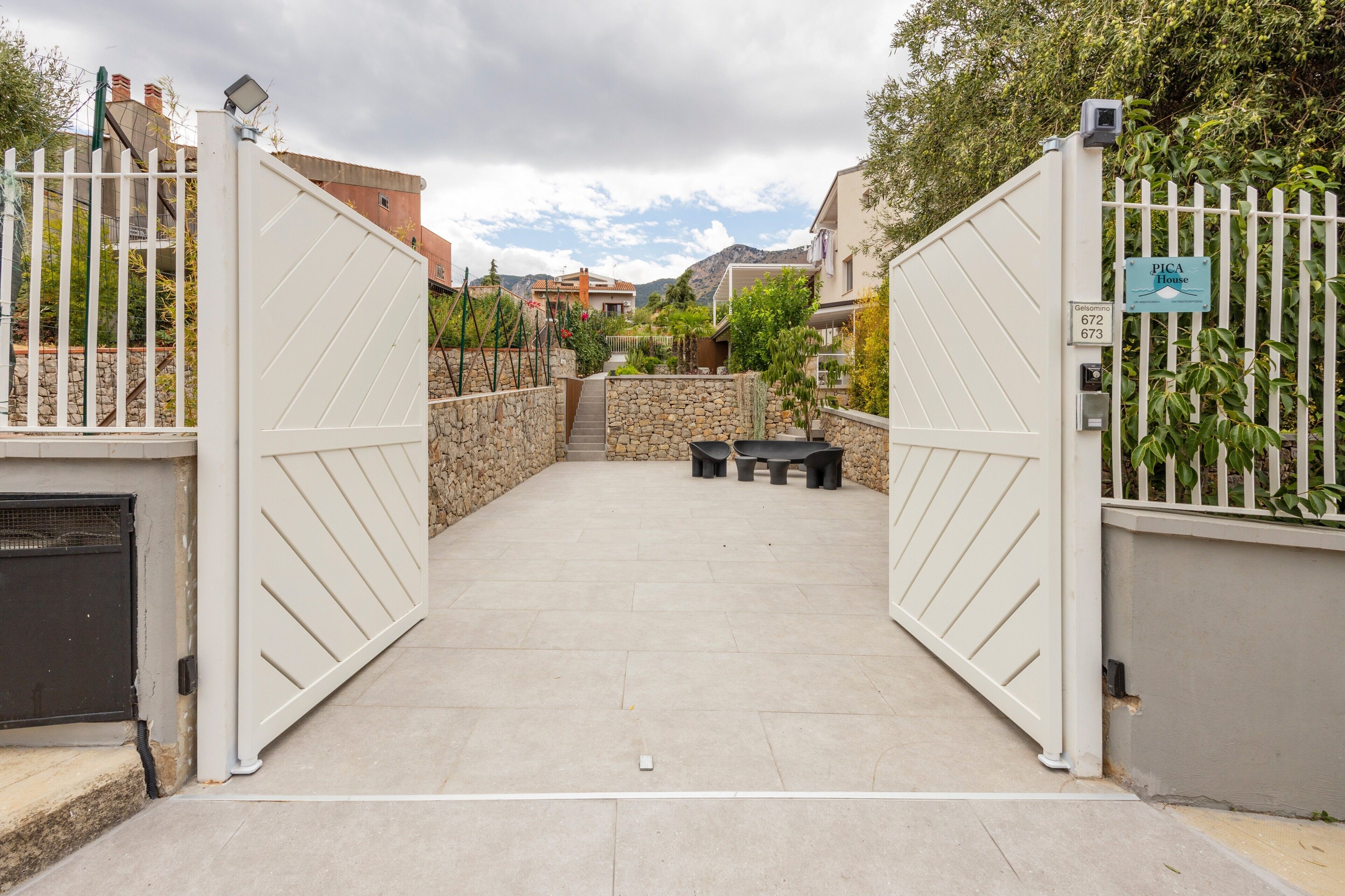 A gated entrance to a property with a stone wall, a sign indicating 'PICA House,' and a view of the surrounding neighborhood.