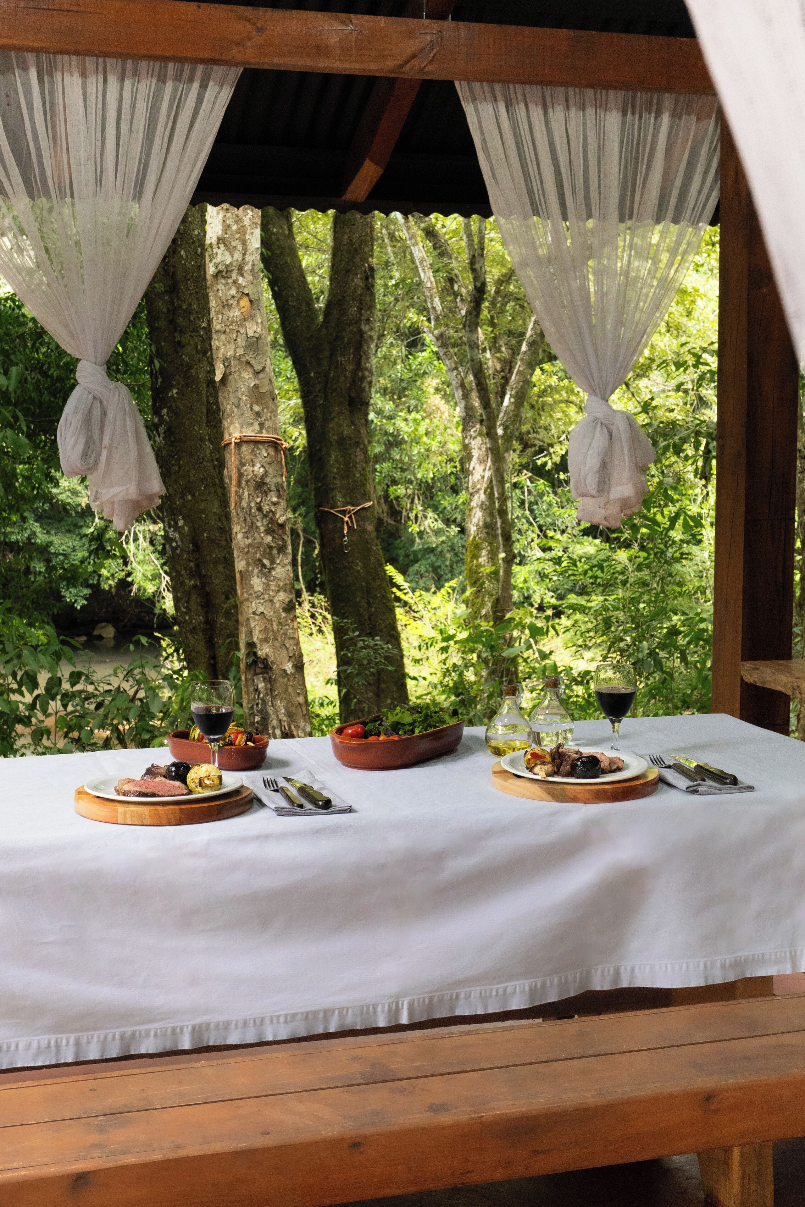 An outdoor dining area with a white tablecloth, wooden benches, and a view of a forested area.