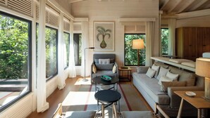 Amplia sala de estar con sofá, sillones, una mesa de centro y ventilador de techo. Grandes ventanales ofrecen abundante luz natural y vistas al exterior.