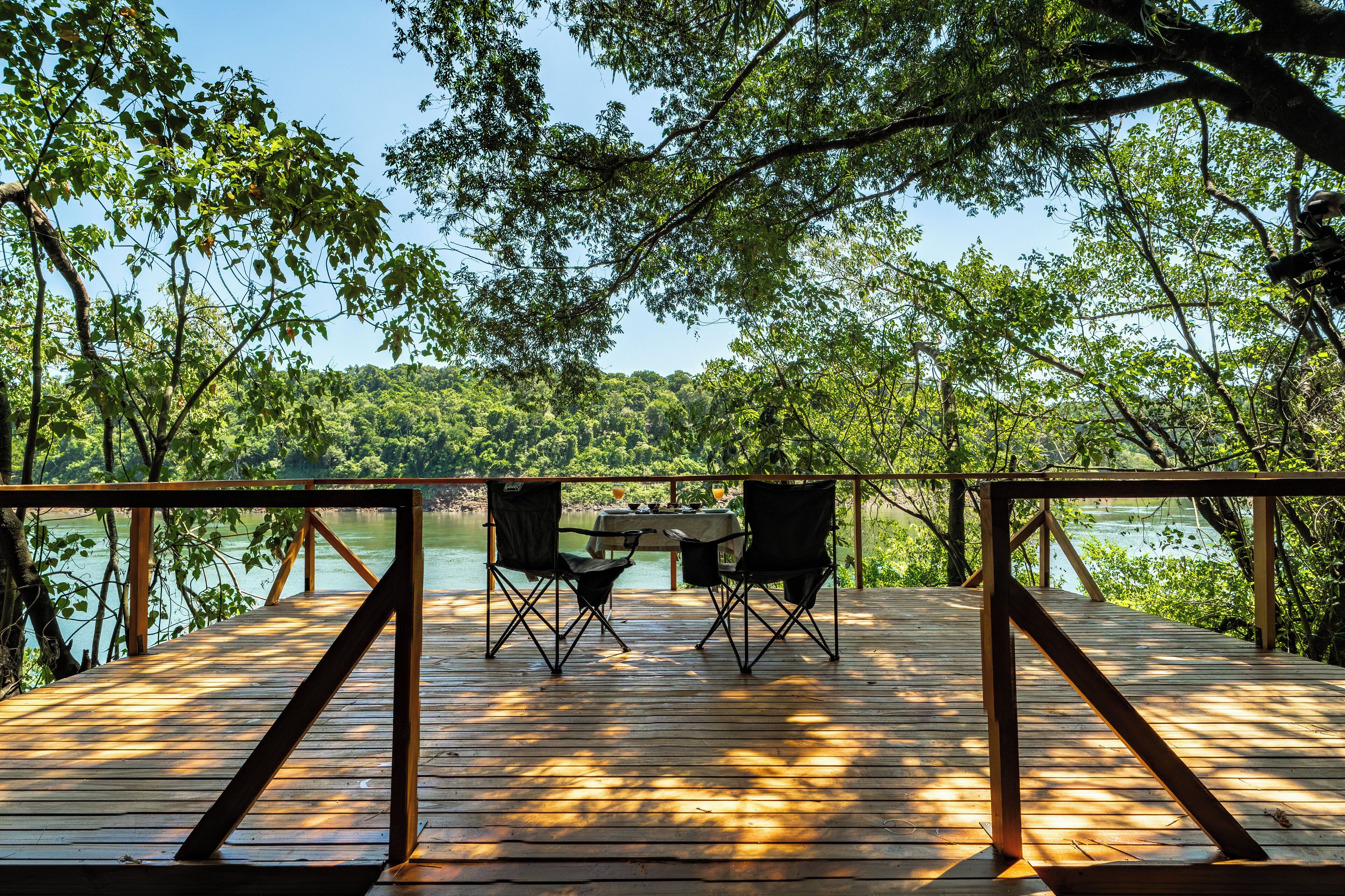 Una terraza de madera con dos sillas y una mesa, con vista a un lago rodeado de árboles.