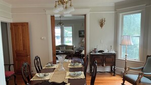 Een eetkamer met een houten tafel voor vier personen, omringd door sierlijke stoelen, een kroonluchter en uitzicht op een woonkamer door een open deur.