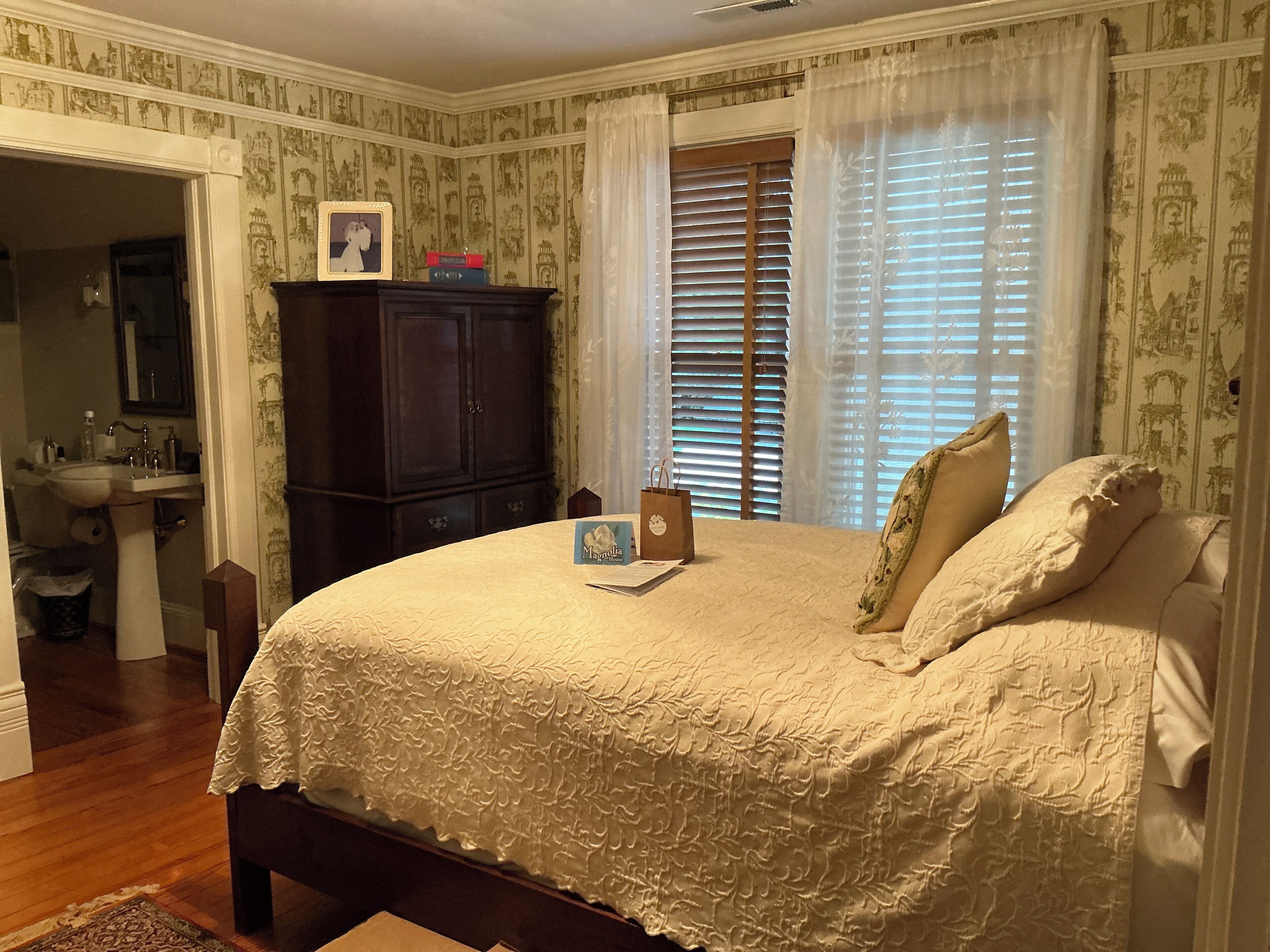 A bedroom with a large bed, a wooden wardrobe, a nightstand, and a window with blinds.